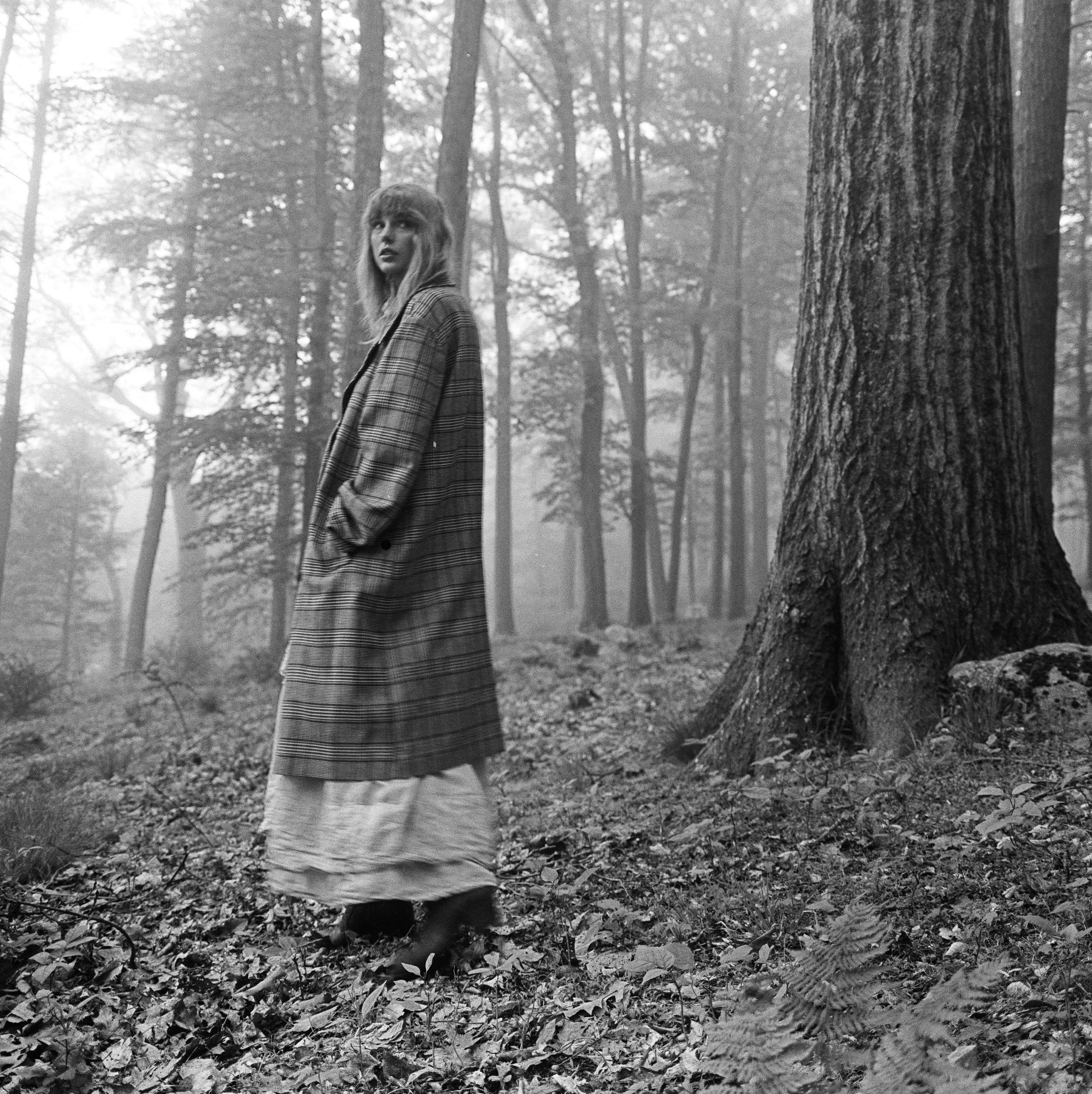 taylor swift looking at the camera while standing in a forest wearing a white dress and large overcoat